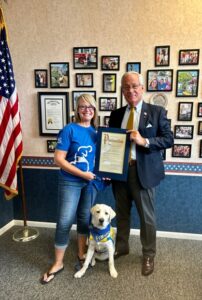 A woman and a man stand inside an office holding a framed proclamation. The woman is smiling, wearing a blue shirt, and holding a dog on a leash. The man is in a suit. A dog sits between them, wearing a vest. There's an American flag and a wall with framed photos in the background.