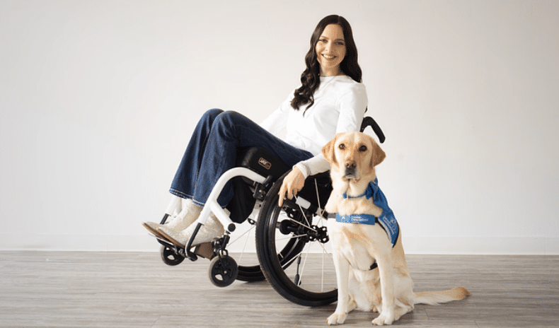 A woman sitting in a wheelchair with a service dog wearing a blue vest beside her.