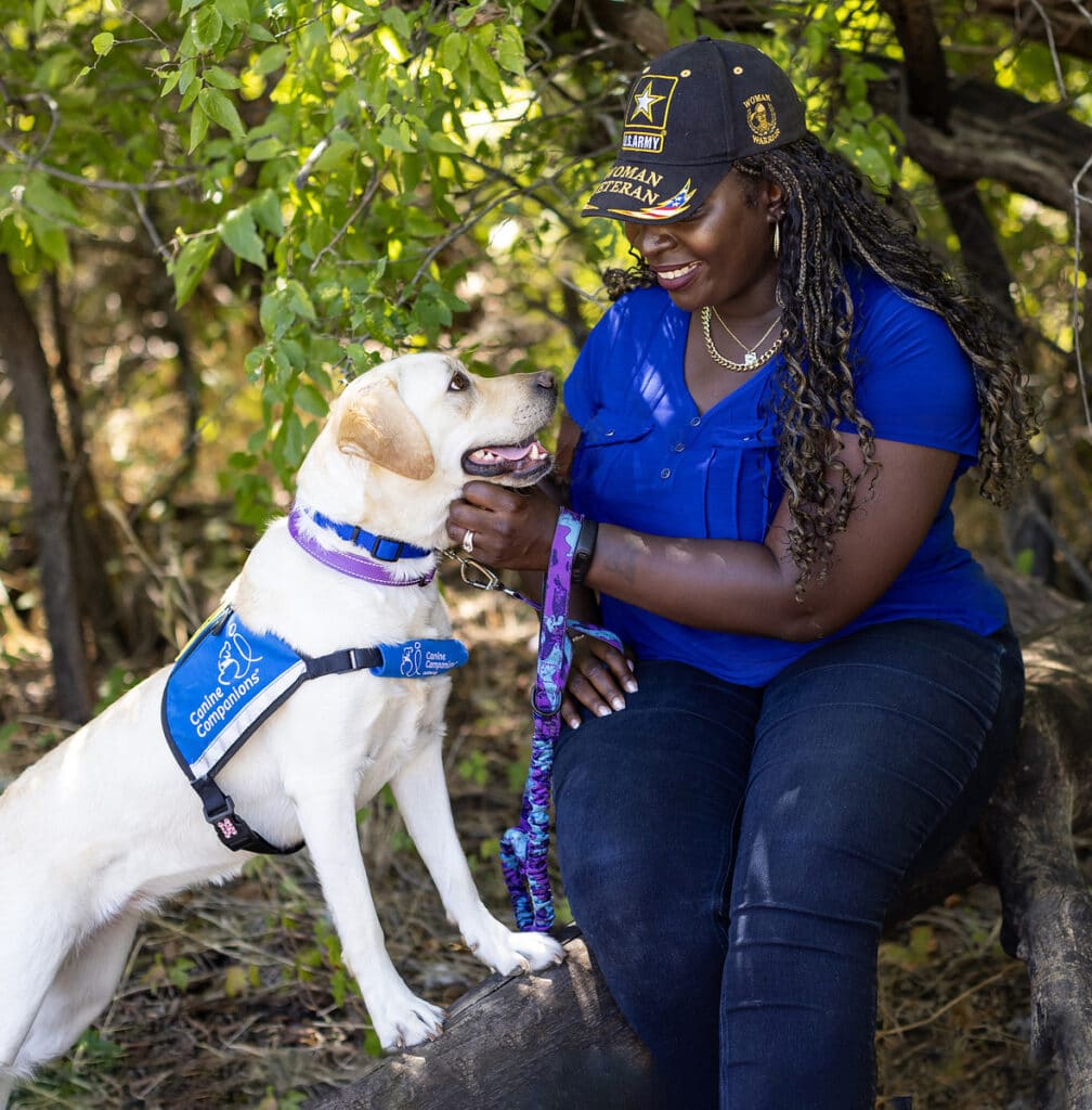 A woman wearing a cap smiles at a Labrador retriever wearing a "Canine Companions" vest, sitting together outdoors with greenery in the background.