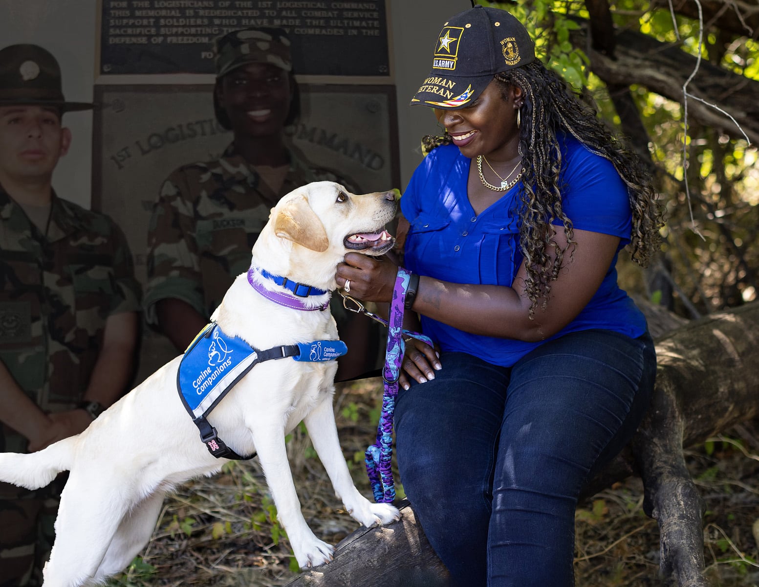 A woman wearing a veteran cap and blue shirt smiling at a Labrador wearing a "Canine Companions" vest.