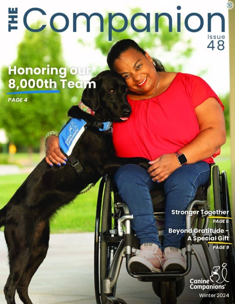 A woman in a wheelchair embraces a black assistance dog outside, smiling. They are featured on the cover of The Companion magazine, issue 48.