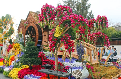 Colorful floral parade float with a rustic water wheel, vibrant flower arrangements, and a yellow butterfly decoration.