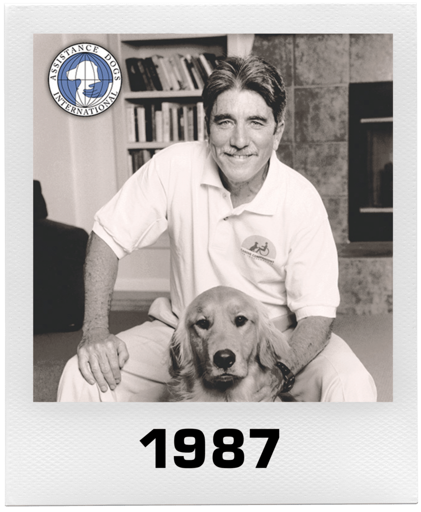 Man smiling with a Golden Retriever, wearing a white Assistance Dogs International polo shirt, dated 1987.