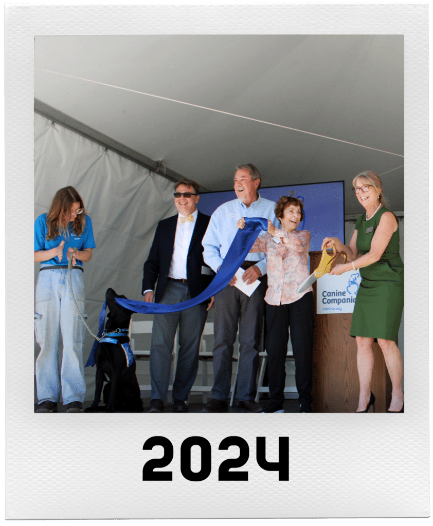 A group of people celebrating with a ribbon-cutting ceremony under a tent, including a service dog wearing a vest.