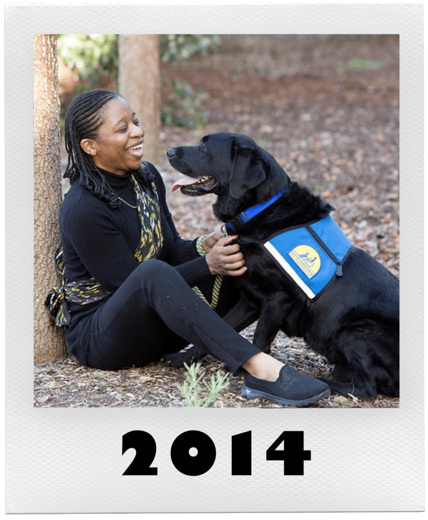 A woman smiling while petting a large black service dog wearing a blue vest in a park setting, with "2014" written at the bottom.