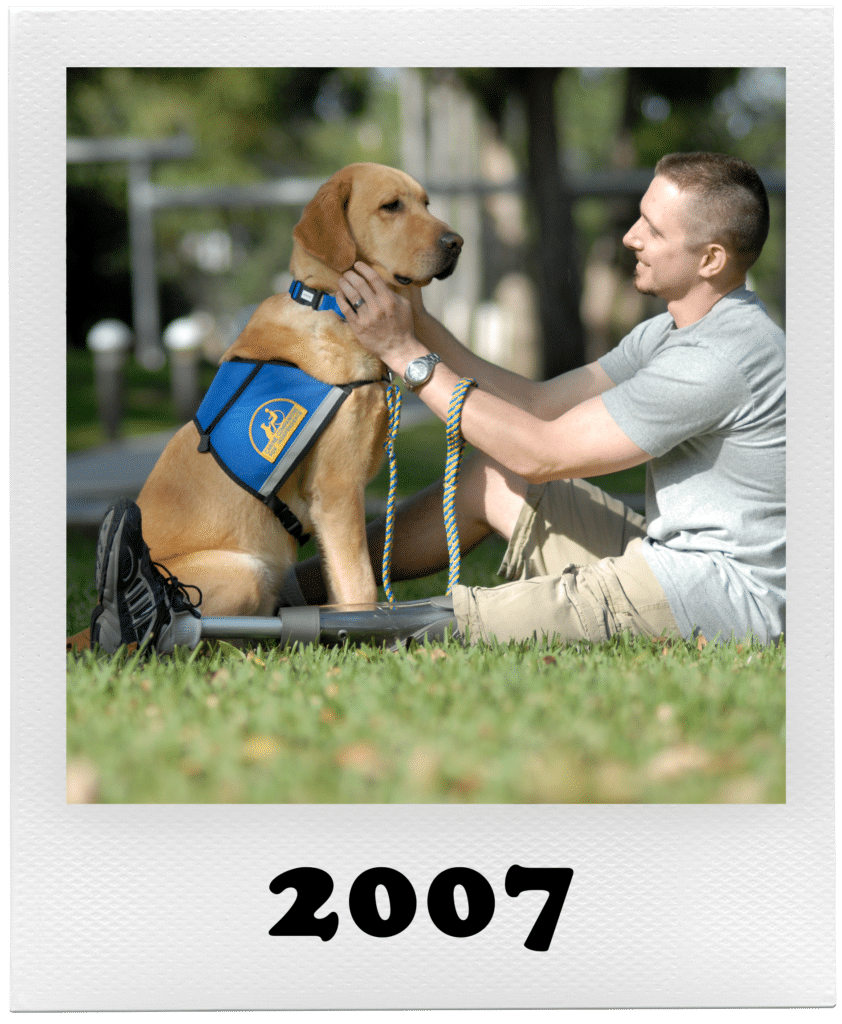 A man in a gray shirt and khaki shorts interacts with a service dog wearing a blue vest, sitting on a grassy area.