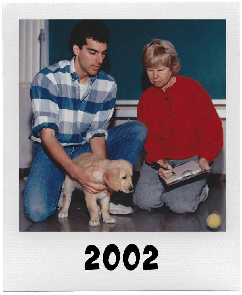 A man and a woman with a golden retriever puppy indoors, with "2002" written at the bottom.