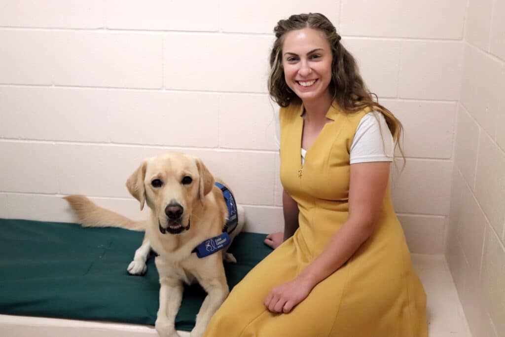 A woman in a yellow dress sitting next to a Labrador retriever wearing a blue service dog vest.