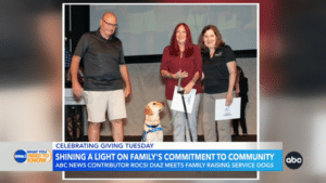 Three people standing on stage with a service dog wearing a blue vest, caption: "Shining a Light on Family's Commitment to Community" on a news segment.