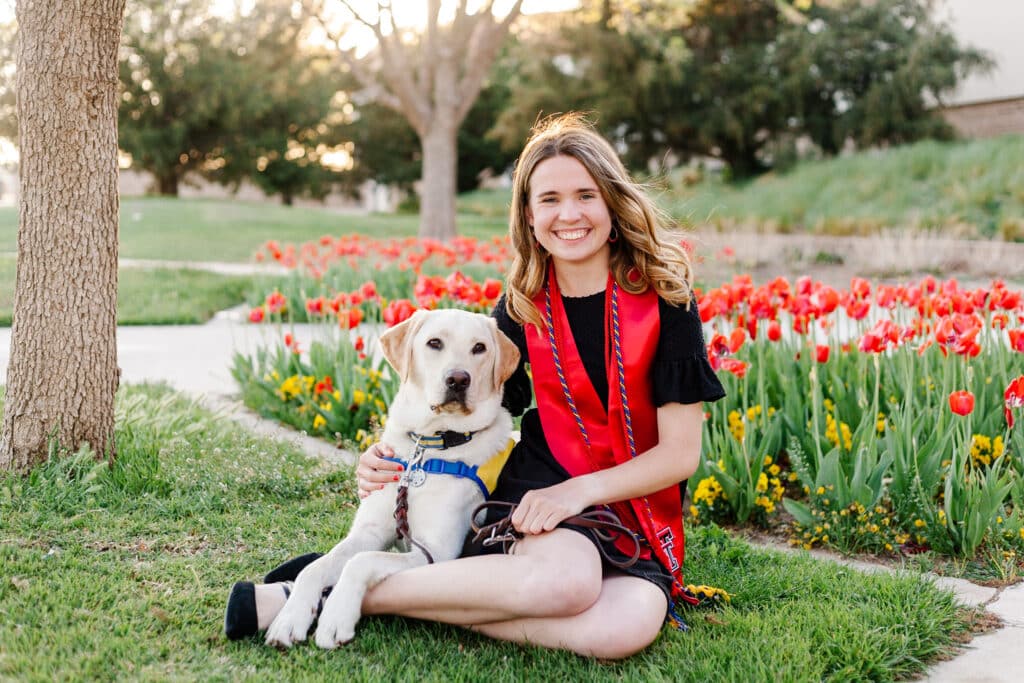 A woman in a graduation outfit sitting on grass with a yellow Labrador Retriever, surrounded by red tulips.