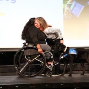 A person in a wheelchair hugs another person while a service dog stands nearby.