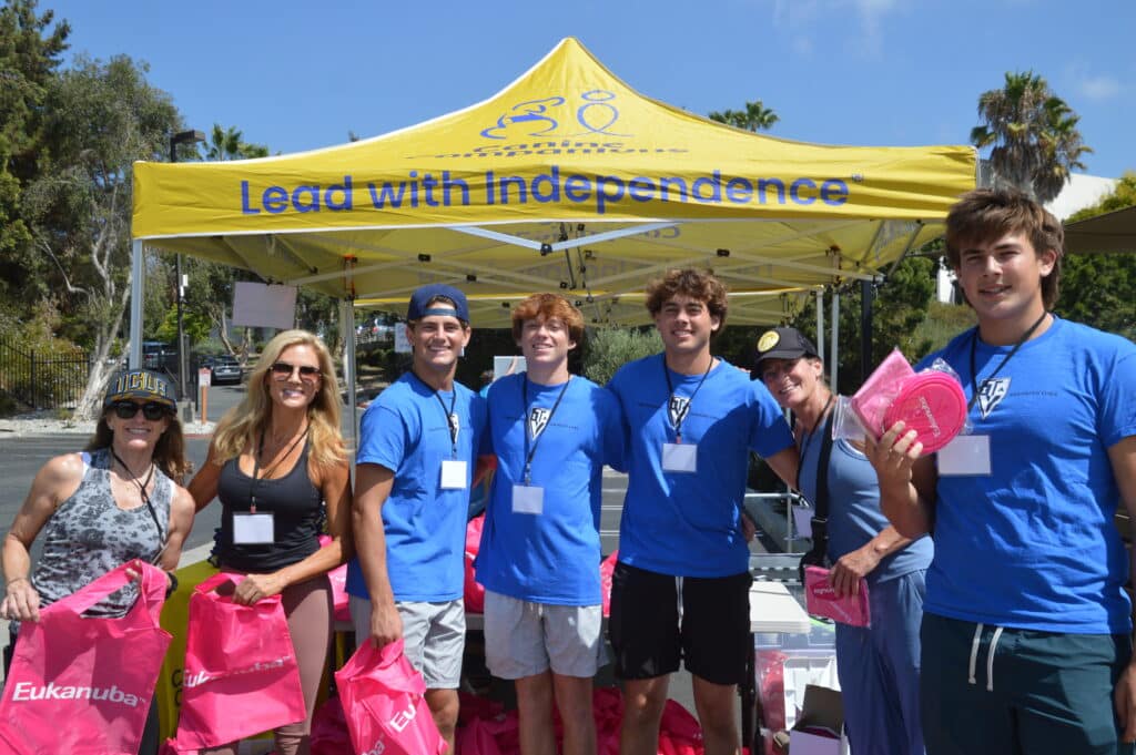 A group of people wearing blue shirts and name tags standing under a yellow tent with the text "Lead with Independence." They are holding pink bags and smiling.