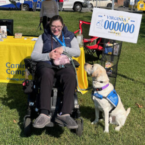 A woman in a wheelchair smiling at a service dog wearing a blue vest, sitting beside a yellow table with a Virginia license plate displayed.