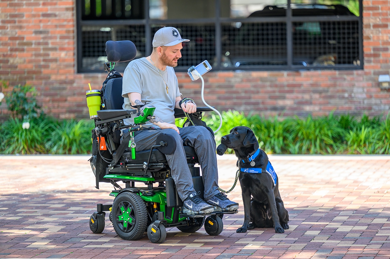 Man in a power wheelchair with a service dog sitting beside him outdoors.