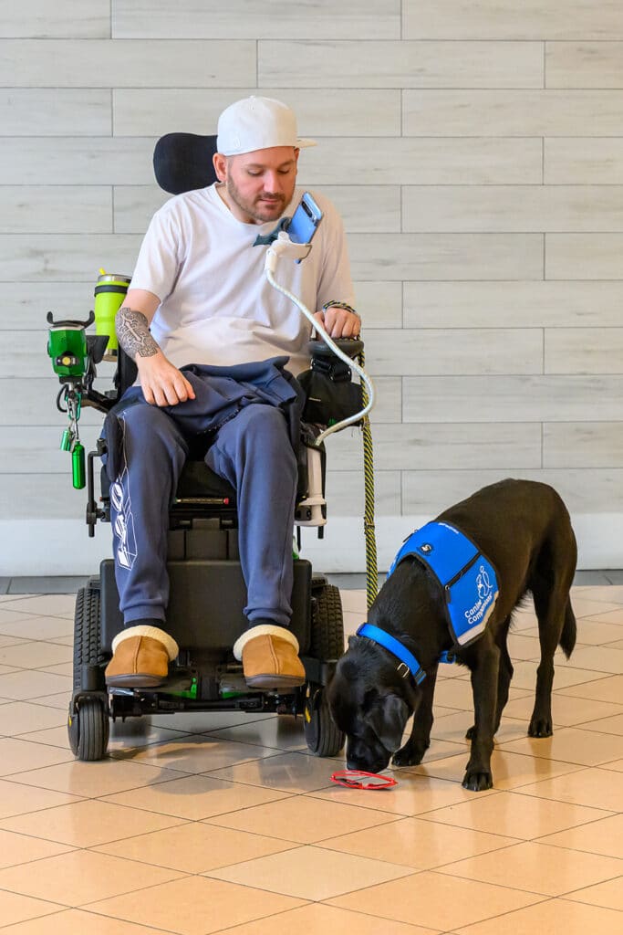 A man in a power wheelchair with a service dog wearing a blue vest, bending down to retrieve a pair of red sunglasses.