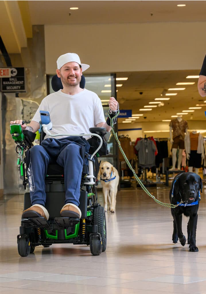 A person in a wheelchair holds a leash, leading a black service dog in a shopping mall.