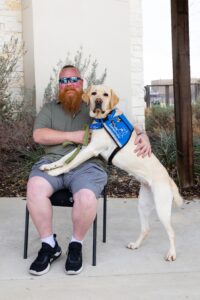 A man sitting on a chair outdoors with a service dog wearing a blue vest standing on hind legs, placing its front paws on the man's lap.