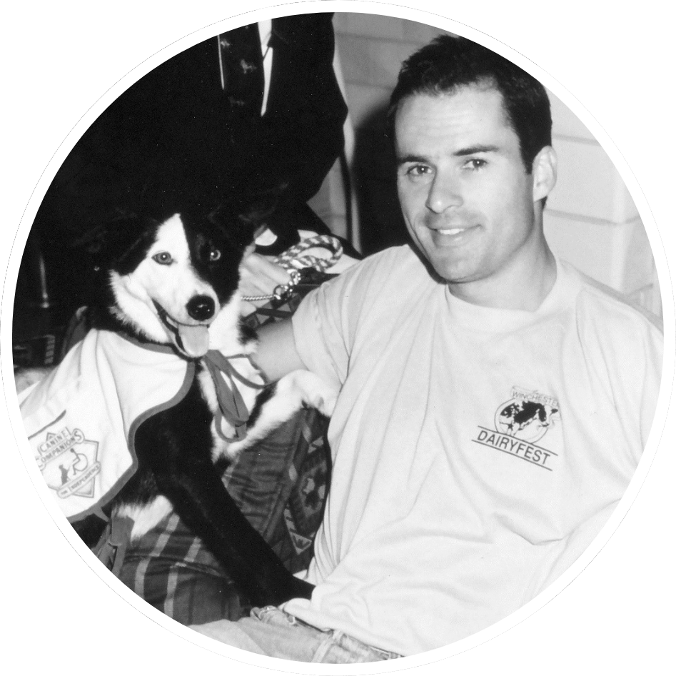 A man smiling and sitting next to a dog wearing a vest.