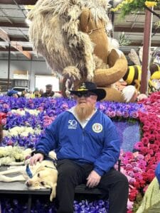 A Canine Companions graduate sits with his service dog on a parade float for the Rose Parade, featuring Lions Club and Canine Companions.