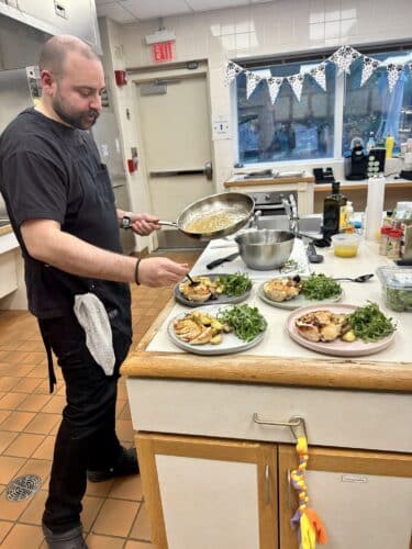 Chef in a kitchen plating grilled chicken, roasted potatoes and arugula on four plates, pouring sauce from a skillet.