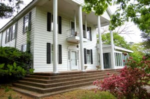 A two-story white house with large columns supporting the porch, black shutters, and a set of stone steps leading up to the front door. There is a glass-enclosed porch on one side and blooming pink flowers in the foreground.