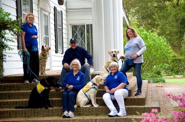 A group of six people, wearing blue shirts, poses on the steps of a white house with green foliage surrounding them. Three dogs, with one wearing a yellow vest, are seated beside the people.