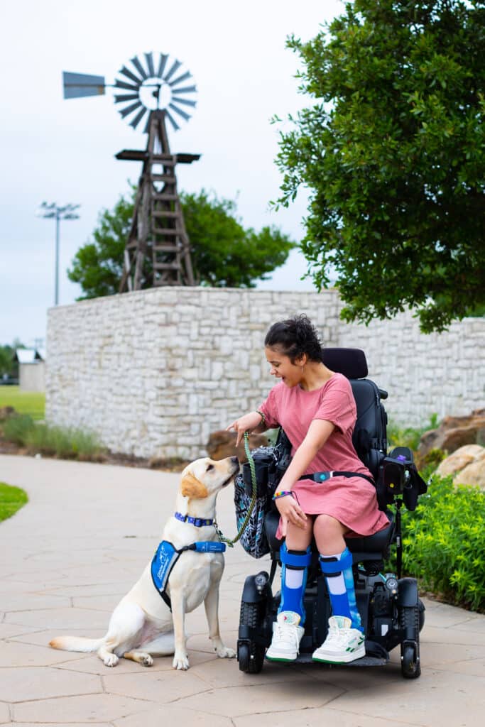 A teen using a power wheelchair outdoors and smiling down at a yellow Labrador/Golden Retriever cross service dog wearing a blue Canine Companions vest.