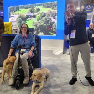 A woman in a blue sweater sits on a mobility scooter with two golden retrievers beside her, while a man in black holds a microphone and gestures towards the crowd in a trade show setting with a large screen behind them.