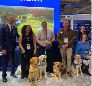 A group of five people stands in front of a display featuring a golf course, with three Labrador Retrievers at their feet. The individuals include a man in a suit, three women, and one man, all smiling. Each person has a name tag, and the dogs are wearing various colored leashes and bandanas.