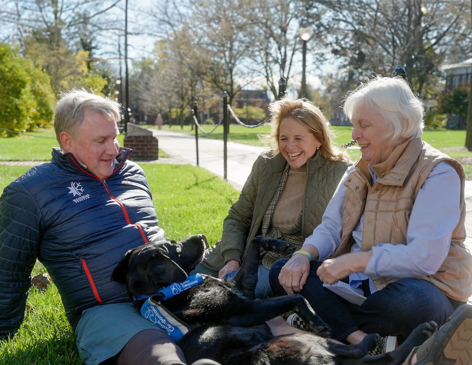 A man and two women sit on the grass in a park, laughing and enjoying time with a black dog lying on its back in front of them. The scene is sunny and features trees in the background.