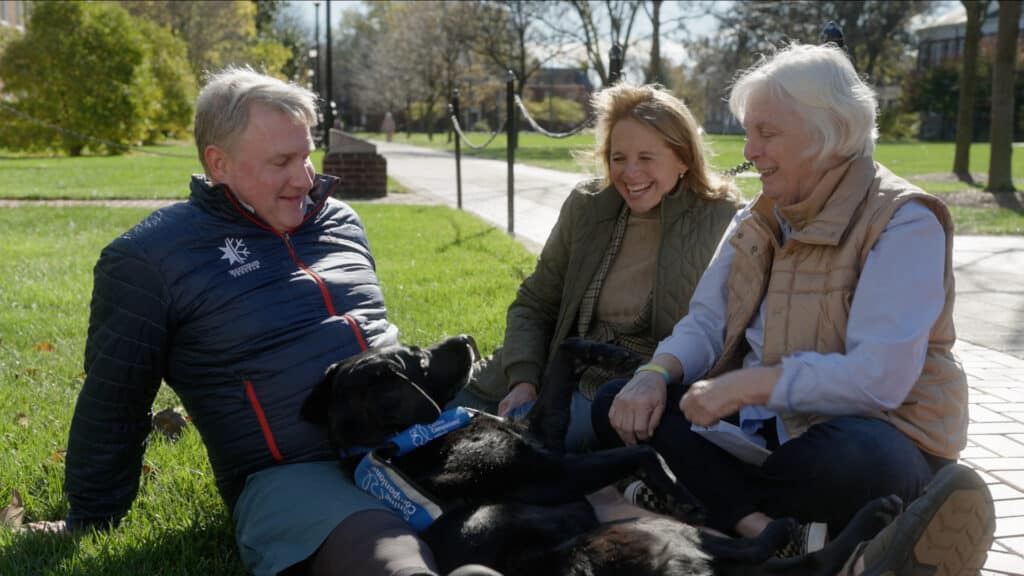 A man in a puffy jacket sits on the grass with two women and a black dog. The group is laughing and enjoying each other's company, with the dog lying on its back playfully. The setting includes trees and a walking path in the background.