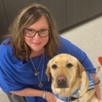 A woman with shoulder-length brown hair, wearing glasses and a blue top, is posing closely with a light-colored Labrador retriever wearing a blue harness. They are indoors, with a speckled gray floor and dark cabinetry in the background.