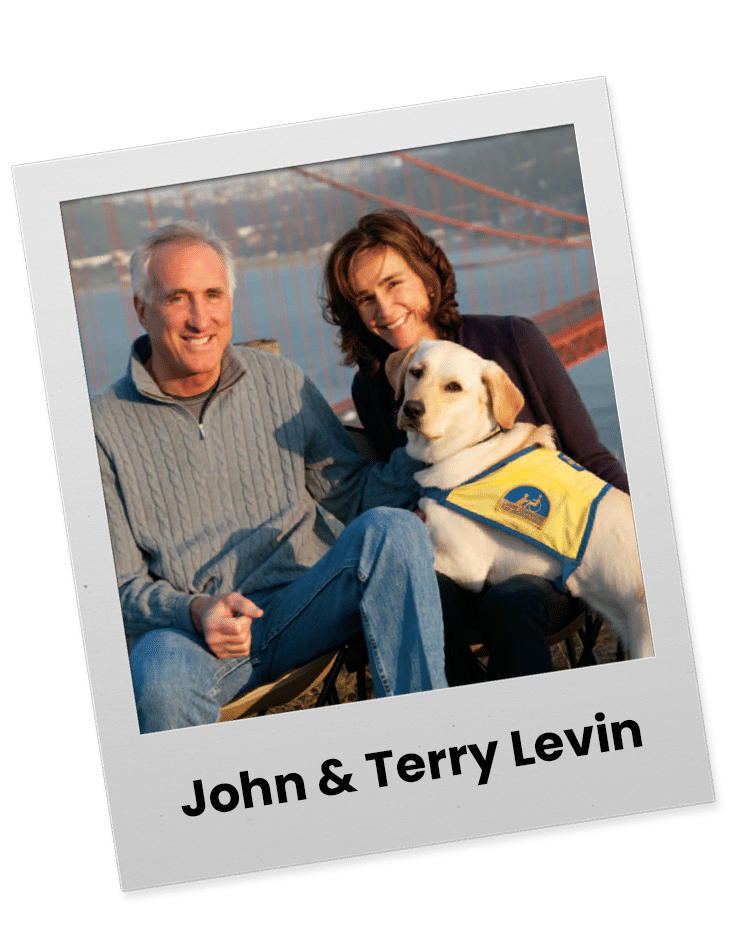 A man and a woman sit together on a bench outdoors with a dog between them, overlooking a body of water and a bridge in the background. The man is wearing a gray sweater and the woman is in a dark top, smiling at the camera. The dog is a yellow labrador wearing a blue vest.