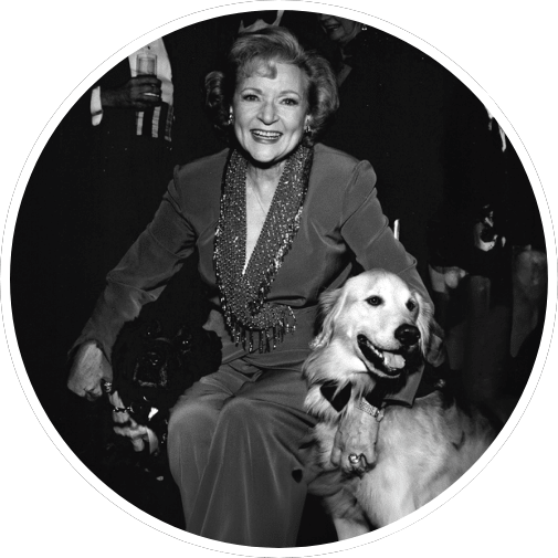 A smiling woman in a sparkling outfit is seated between two dogs, a black dog and a golden retriever, in a black and white photograph.