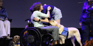 A woman in a wheelchair hugs a young girl while a yellow Labrador wearing a "Canine Companions" vest sits beside them. Another woman stands nearby, and the setting appears to be a stage with dim lighting.