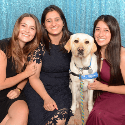 Three women pose together with a Labrador retriever in front of a shiny blue backdrop, smiling and enjoying the moment.