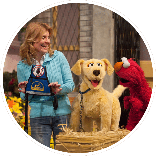 A woman in a blue sweater smiles while holding a framed award next to a large, animated golden dog puppet and a red furry character resembling a Muppet in a colorful outdoor setting with flowers and hay bales.