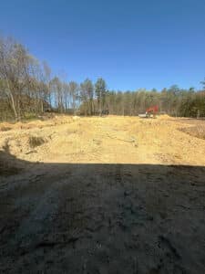 A cleared construction site with uneven earth and mounds of sand under a clear blue sky; a red excavator is visible in the background among sparse trees.