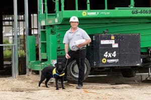 A person wearing a gray shirt and hard hat stands next to a black dog, dressed in a yellow vest, in front of a green construction lift and equipment, with construction site elements visible in the background.
