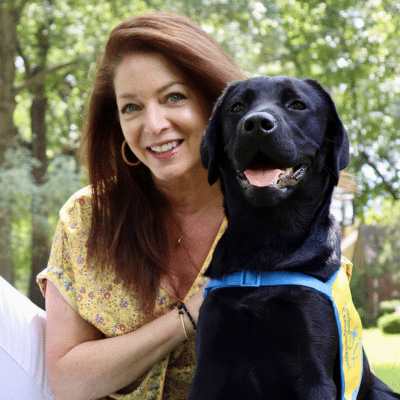 A woman with long brown hair smiles as she sits next to a black Labrador retriever, both outdoors in a green setting. The dog wears a blue harness with a yellow service dog vest, and they appear to be enjoying each other's company.