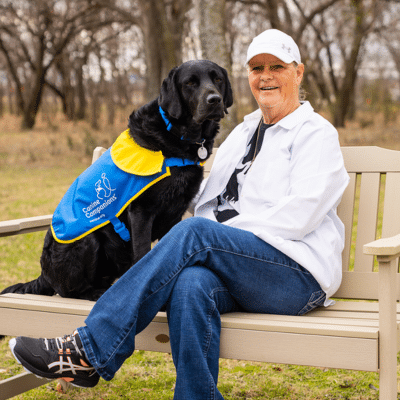 A woman in a white jacket and a cap sits on a bench next to a black Labrador retriever wearing a blue and yellow service dog vest in a park setting with trees in the background.