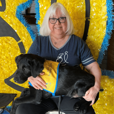 A woman with shoulder-length white hair and glasses holds a black dog wearing a service vest, sitting against a bright yellow and blue background decorated with a playful design.