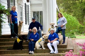 A group of five people in blue shirts and two dogs, one black and one yellow, are gathered on the steps of a white house surrounded by greenery. Two women stand on the left, while three others sit on the steps with one person on each side of two dogs in training harnesses.