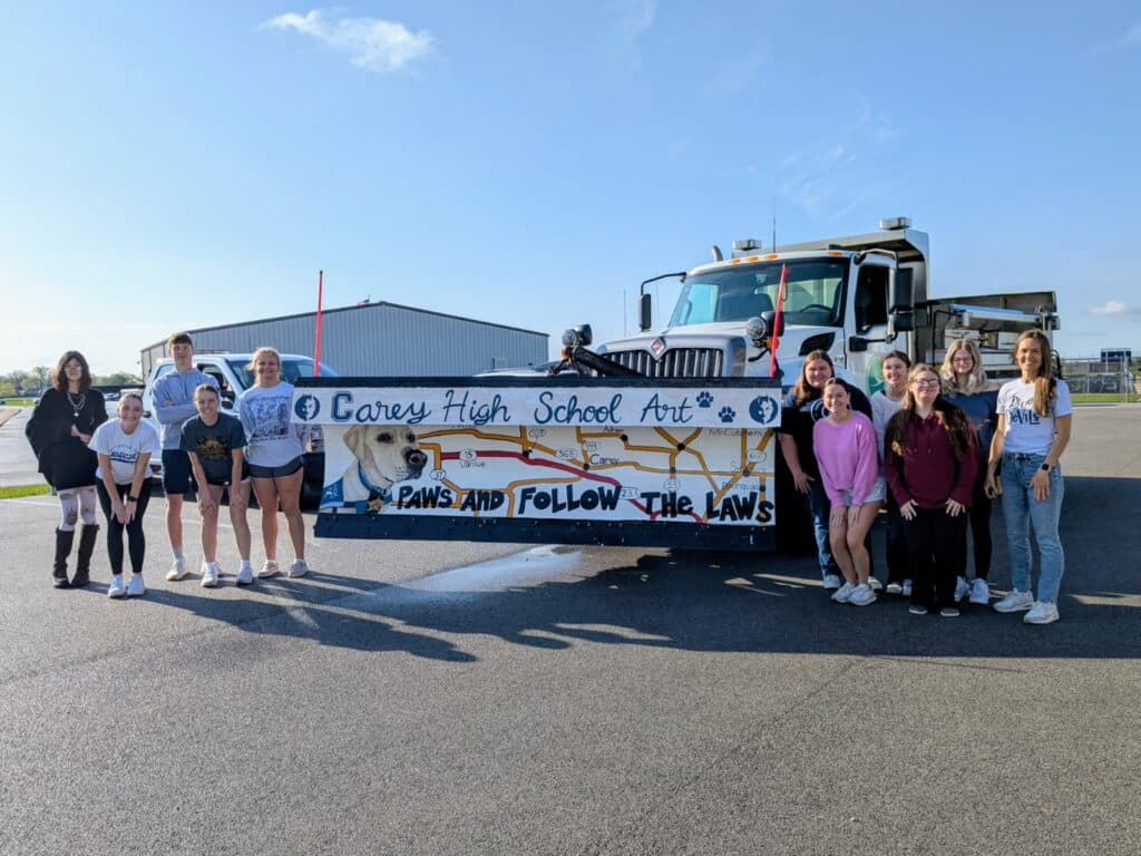 Students and a teacher pose in front of a decorated vehicle displaying the logo 'Carey High School Art' and the phrase 'Paws and Follow the Laws', with a dog illustration and a map-like design. The setting is an outdoor area with a clear blue sky.