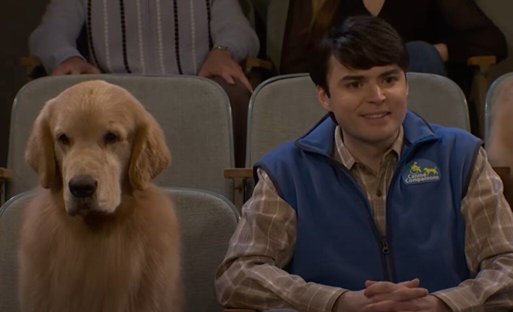A golden retriever sitting next to a young man in a blue vest with a logo, both seated in a theater setting with audience members visible in the background.