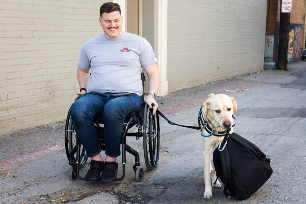 A man in a wheelchair smiles while holding the leash of a yellow dog, which is carrying a black bag in its mouth, on a street with a brick wall in the background.