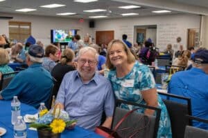 A smiling elderly man with glasses wearing a blue striped shirt is seated at a table, next to a woman in a blue patterned dress with a name tag that says "Linda." They are at a social gathering surrounded by other attendees and decorated tables with flowers and bottles of water.