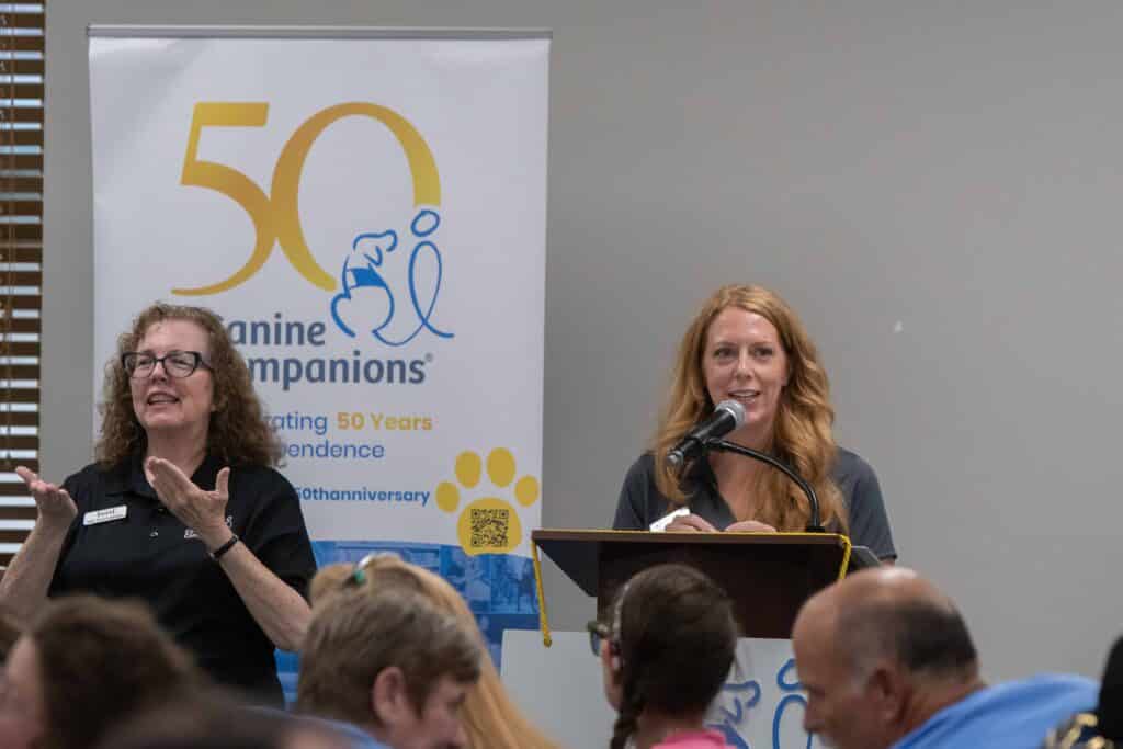 A woman with long, wavy red hair speaks into a microphone at a podium, while another woman with curly brown hair gestures beside her. They are at an event celebrating 50 years of Canine Companions, indicated by a banner in the background.