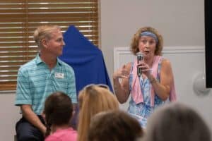 A woman with curly hair, wearing a blue dress and a pink wrap, speaks into a microphone while gesturing with her hands. Beside her, a man in a teal striped polo shirt listens. The setting appears to be a casual indoor event with wooden blinds in the background.