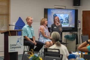 Three speakers seated on chairs during a presentation at an event celebrating the 25th anniversary of the Southeast Region SeaWorld Campus; a woman with red hair speaks into a microphone while the two seated next to her listen, with a Canine Companions logo displayed on a podium in front.